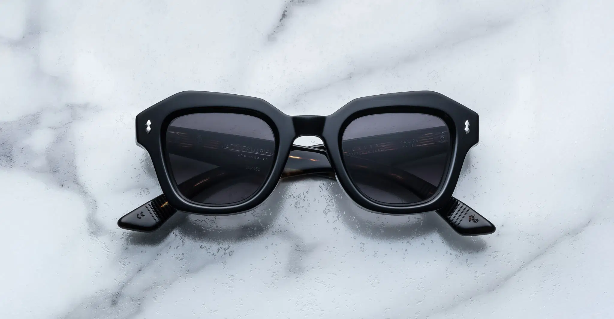 A pair of Jacques Marie Mage Eichler black, thick-framed sunglasses with grey lenses rests on a white marble surface with grey veins, with faint brand details visible on the inside of the left lens.