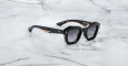 A pair of Jacques Marie Mage Eichler black, thick-framed sunglasses with grey lenses sits on a white marble surface with grey veins