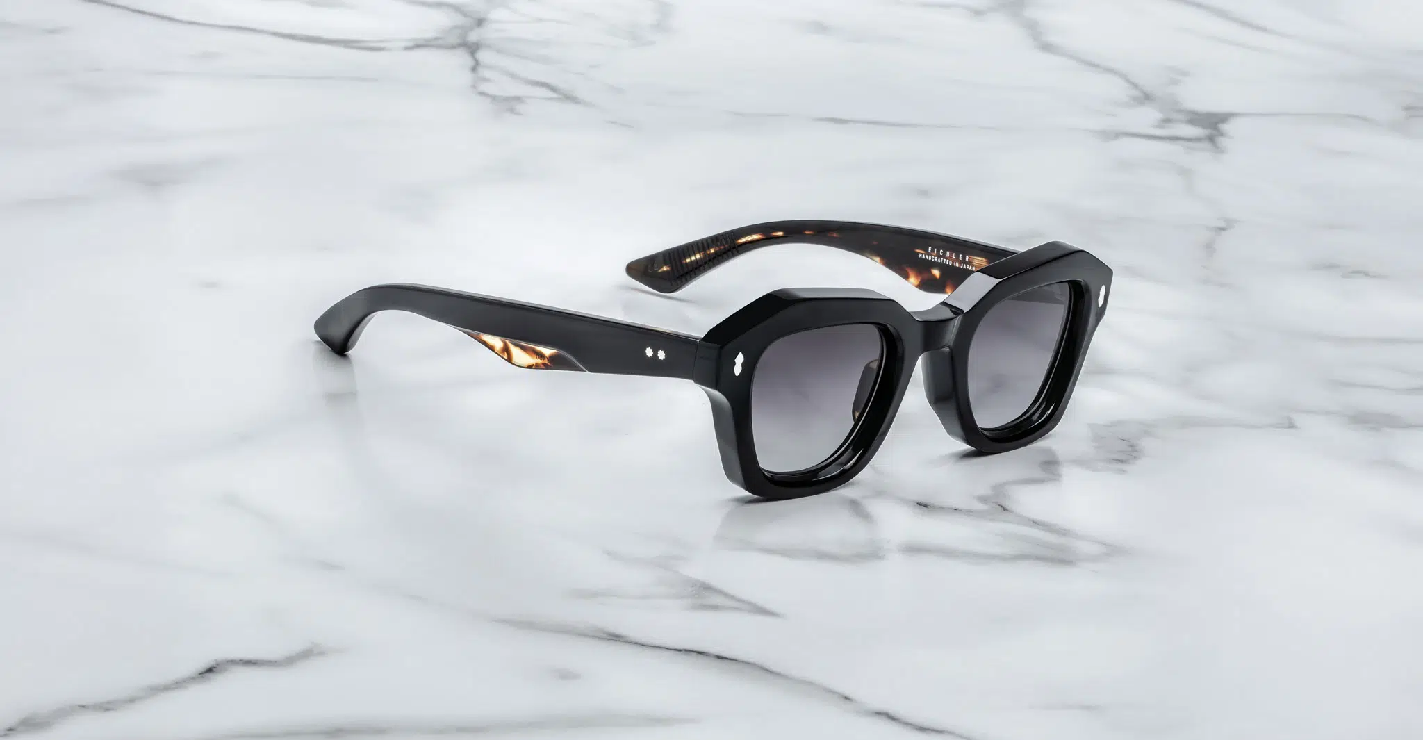 A pair of Jacques Marie Mage Eichler black, thick-framed sunglasses with grey lenses sits on a white marble surface with grey veins