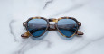 The Jacques Marie Mage Hatfield sunglasses, featuring brown tortoiseshell frames and blue-tinted lenses, are displayed on a white marble surface.