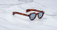 The Jacques Marie Mage Kellerman sunglasses, featuring round tortoiseshell frames and dark lenses, rest at an angle on a white marble surface, showcasing both their side profile and front view.