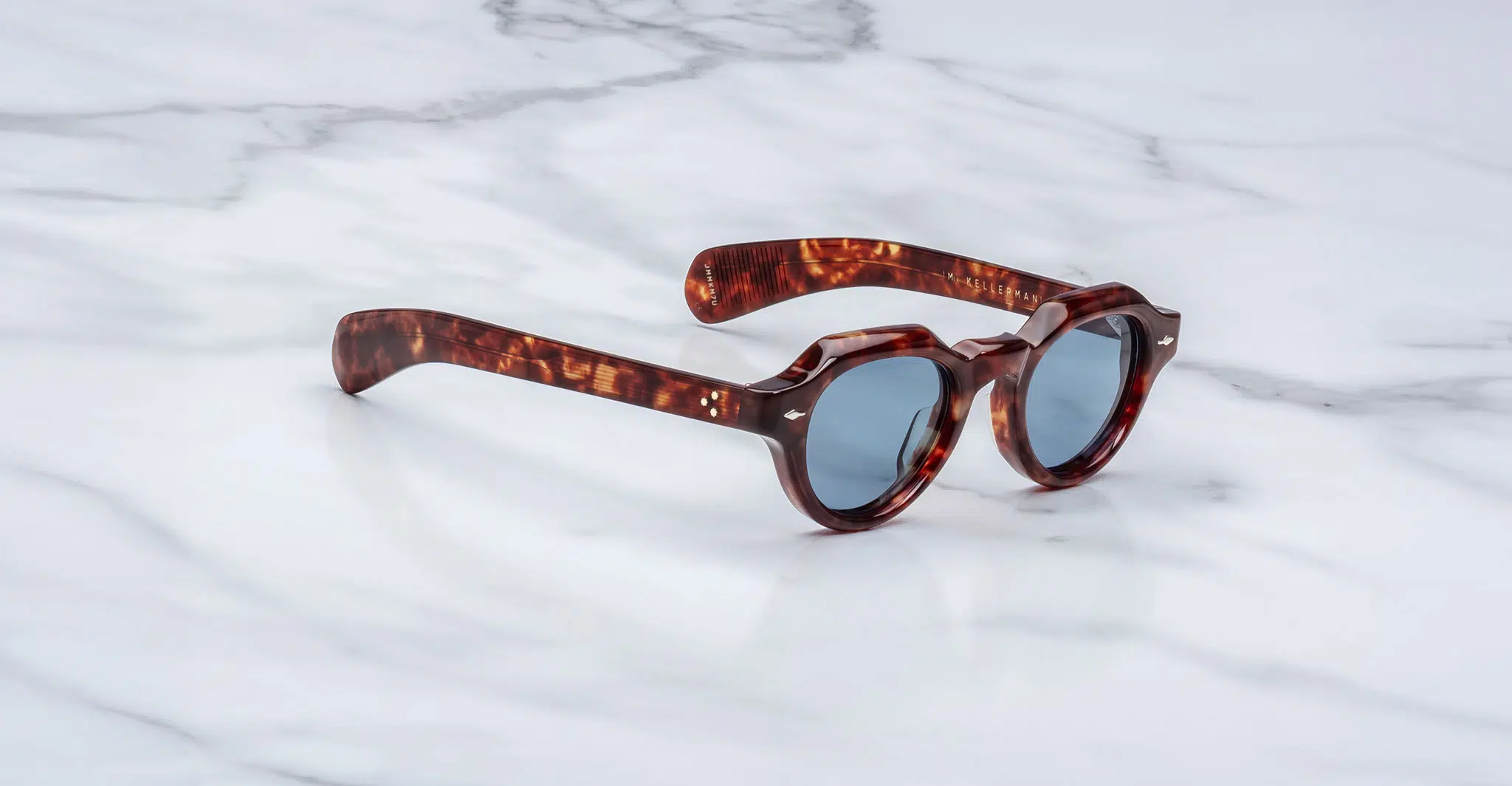 Jacques-Marie-Mage-Kellerman-Breccia-JMMKM-7U-3-4-Dan-Deutsch The Jacques Marie Mage Kellerman sunglasses, featuring round tortoiseshell frames and dark lenses, rest at an angle on a white marble surface, showcasing both their side profile and front view.