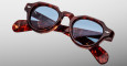 The Jacques Marie Mage Kellerman eyeglasses feature round, thick tortoiseshell frames with blue tinted lenses, shown against a plain light gray background with the glossy arms crossed.