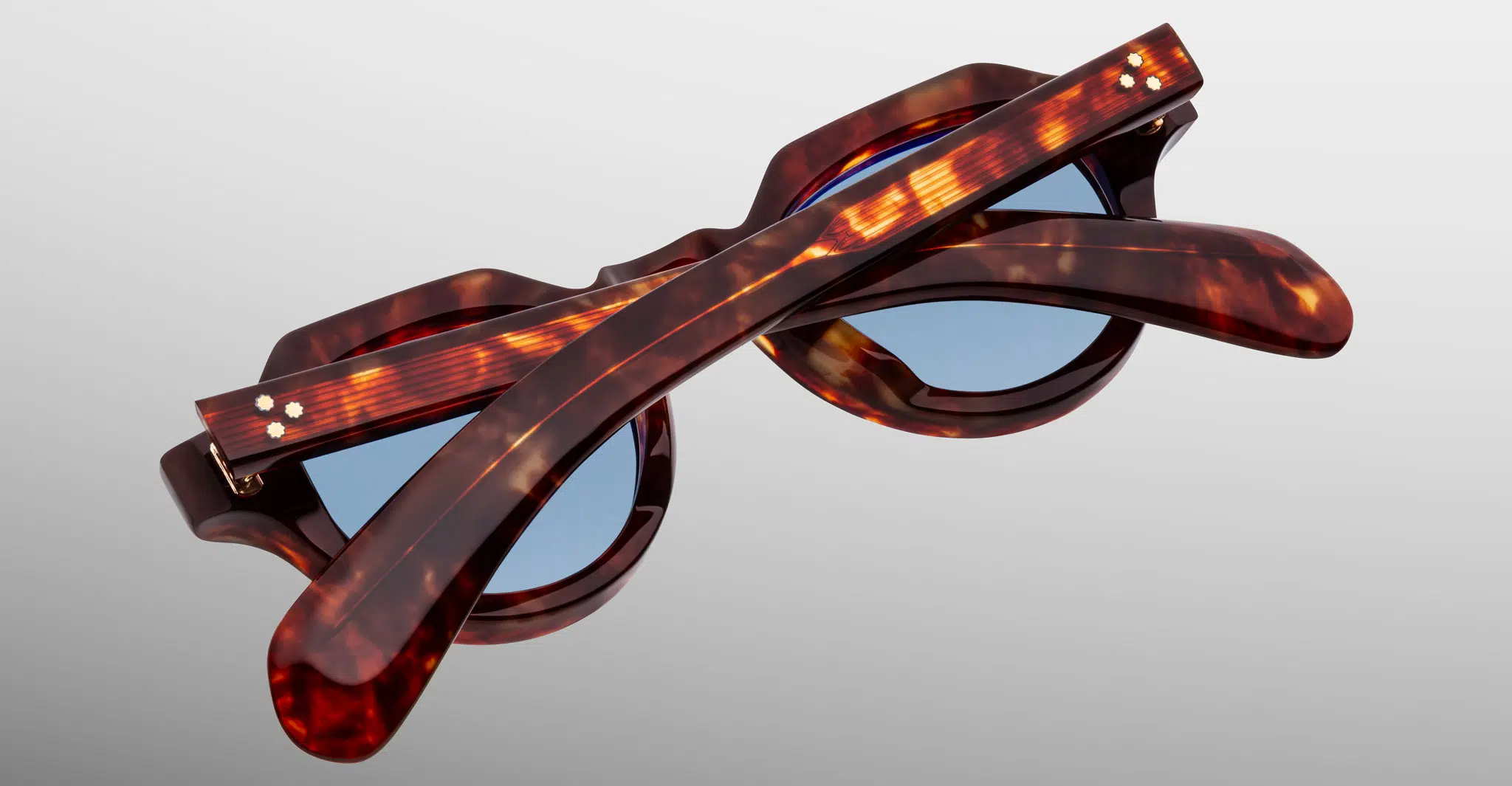 Jacques-Marie-Mage-Kellerman-Breccia-JMMKM-7U-Detail3-Dan-Deutsch The Jacques Marie Mage Kellerman sunglasses, featuring tortoiseshell frames and blue lenses, are shown folded shut on a light gray background in a top-down view that highlights their elegant design.