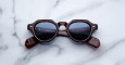 Jacques Marie Mage Kellerman round tortoiseshell sunglasses with blue-tinted lenses rest upside down on a white marble surface, temples folded neatly.