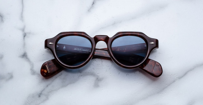 Jacques Marie Mage Kellerman round tortoiseshell sunglasses with blue-tinted lenses rest upside down on a white marble surface, temples folded neatly.