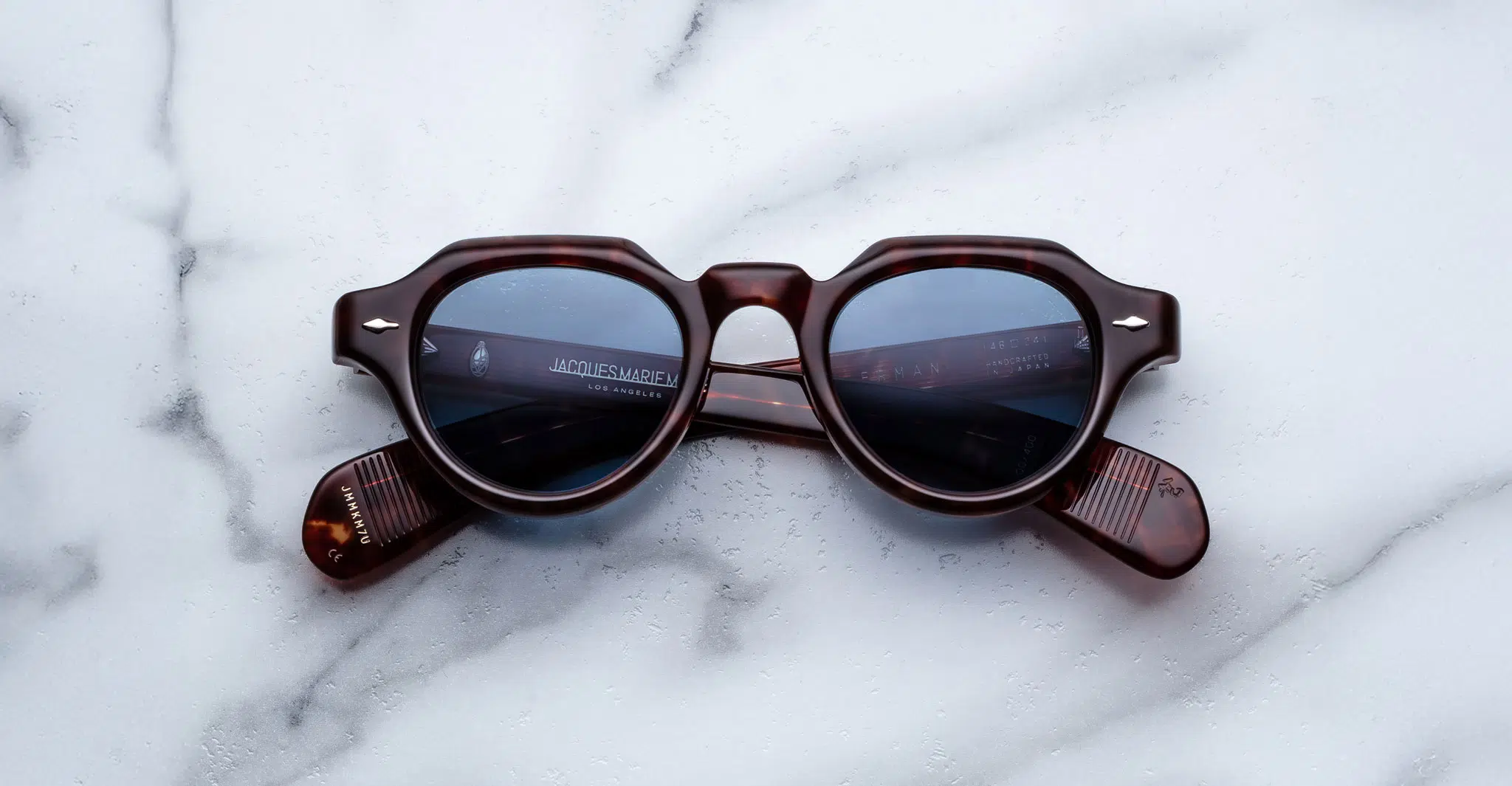 Jacques-Marie-Mage-Kellerman-Breccia-JMMKM-7U-Front-Dan-Deutsch Jacques Marie Mage Kellerman round tortoiseshell sunglasses with blue-tinted lenses rest upside down on a white marble surface, temples folded neatly.
