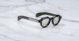 A pair of Jacques Marie Mage Kellerman sunglasses with black round lenses, silver arm details, and a green inner arm featuring gold text, resting on a white marble surface.