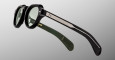 A close-up of Jacques Marie Mage Kellerman black round sunglasses with green lenses, metallic temple detailing, and an olive green inner arm with gold branding, set against a plain gray background.