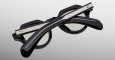 The Jacques Marie Mage Kellerman sunglasses, black and round with green-tinted lenses, are shown folded closed with crossed arms in front of the frames on a light gray background.