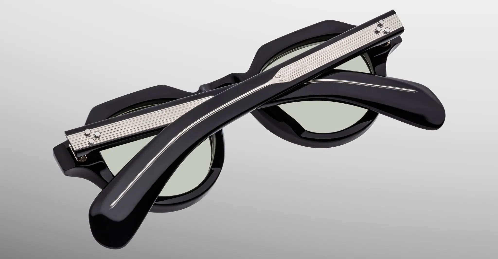 The Jacques Marie Mage Kellerman sunglasses, black and round with green-tinted lenses, are shown folded closed with crossed arms in front of the frames on a light gray background.