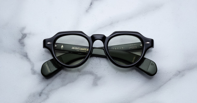A pair of Jacques Marie Mage Kellerman thick black round eyeglasses with dark tinted lenses rests on a white marble surface, folded arms revealing text inside the frames.