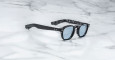 The Jacques Marie Mage Zephirin sunglasses feature blue-tinted lenses and a modern black frame with a black-and-white marbled pattern on the inner arms, shown resting on a white marble surface.