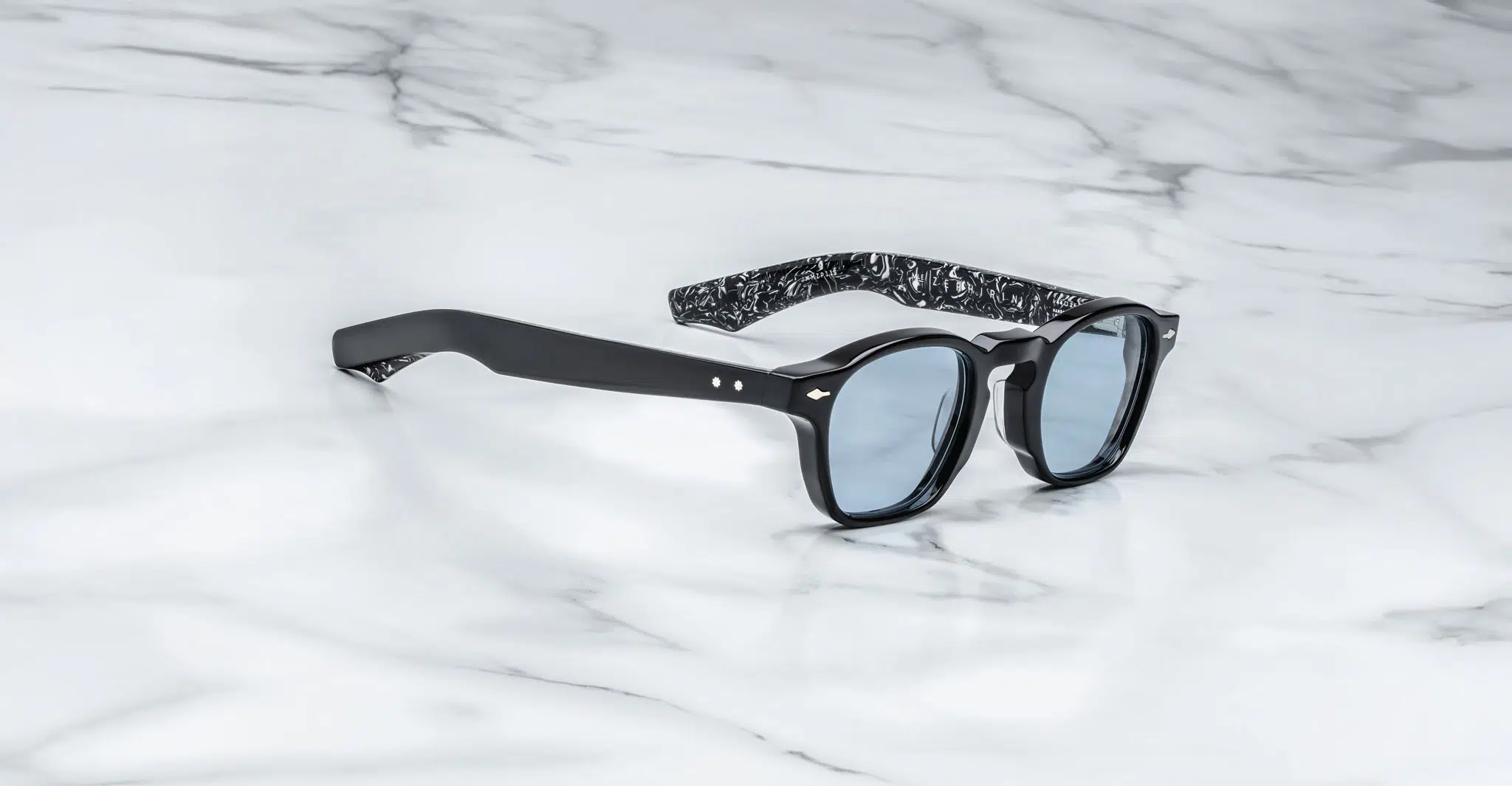 The Jacques Marie Mage Zephirin sunglasses feature blue-tinted lenses and a modern black frame with a black-and-white marbled pattern on the inner arms, shown resting on a white marble surface.
