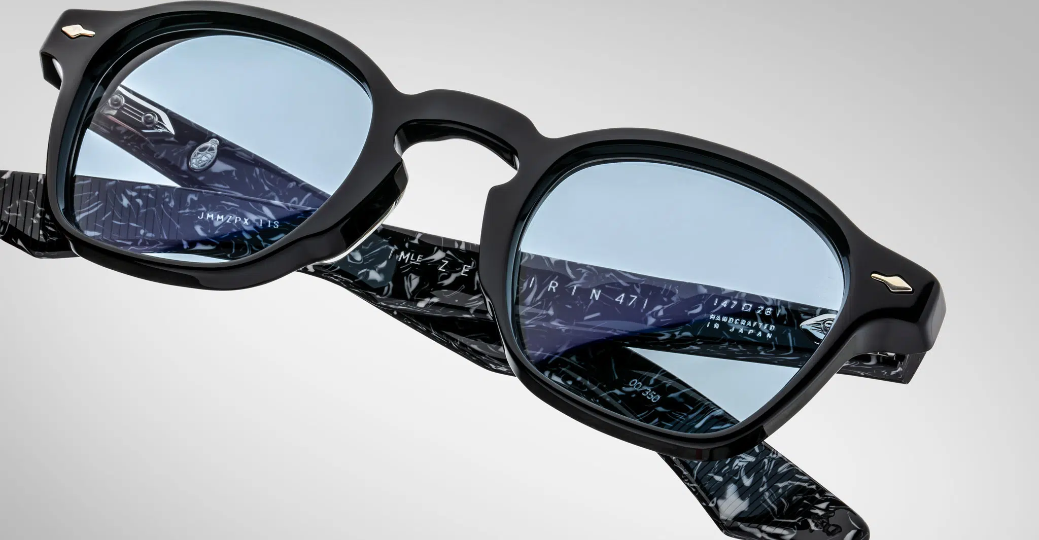 Close-up of Jacques Marie Mage Zephirin eyeglasses with black frames and blue-tinted lenses. The temple interiors display a black and white marbled pattern with engraved text, set against a light gray background.