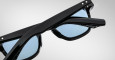 Close-up of Jacques Marie Mage Zephirin black sunglasses with blue-tinted lenses, partially folded, on a plain light gray background.