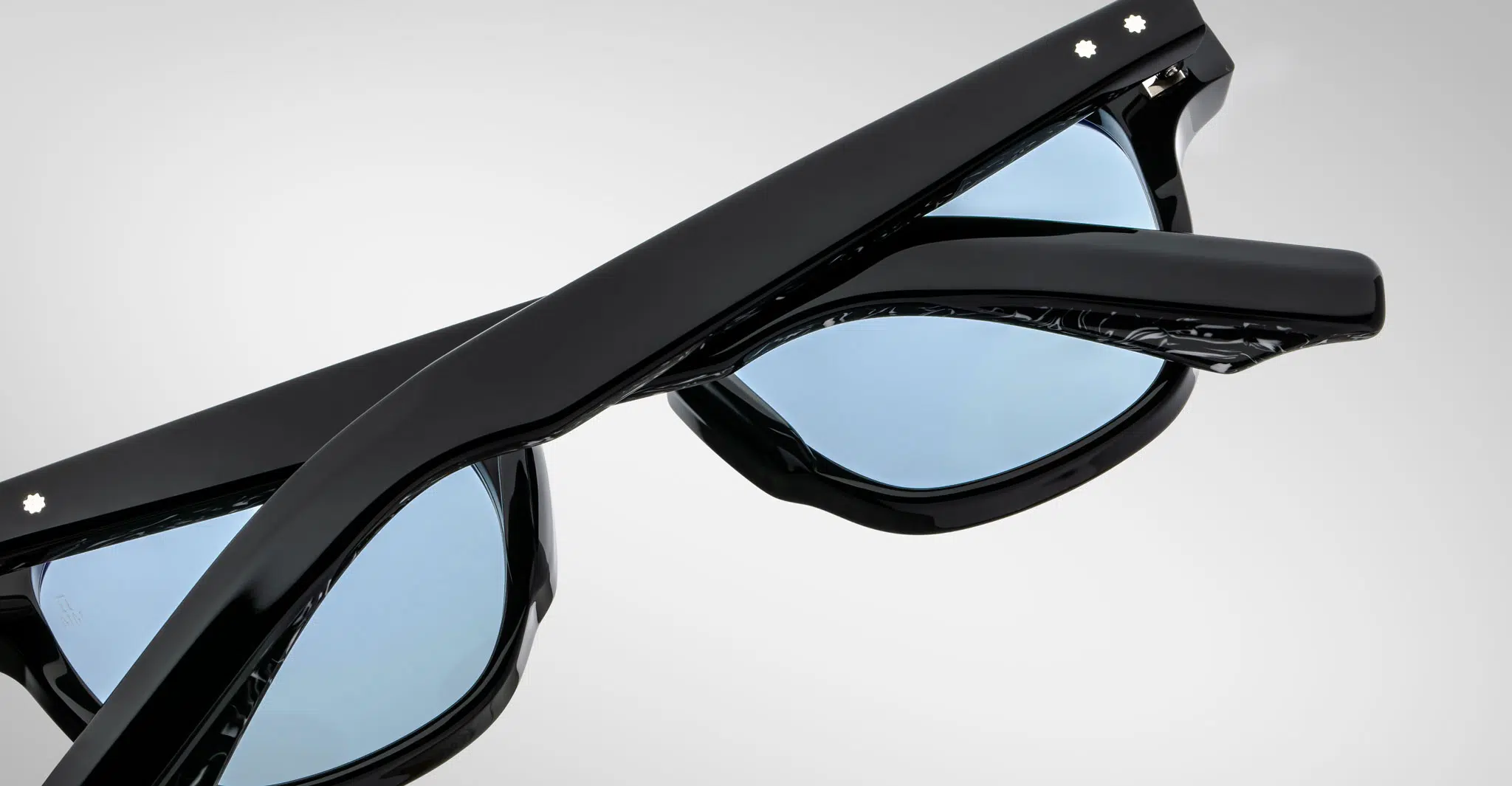 Close-up of Jacques Marie Mage Zephirin black sunglasses with blue-tinted lenses, partially folded, on a plain light gray background.
