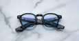 The Jacques Marie Mage Zephirin features black frames, blue-tinted lenses, and marbled black-patterned arms with text on the inside, shown displayed on a white marble surface.