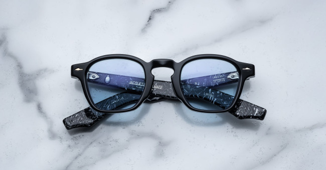 The Jacques Marie Mage Zephirin features black frames, blue-tinted lenses, and marbled black-patterned arms with text on the inside, shown displayed on a white marble surface.