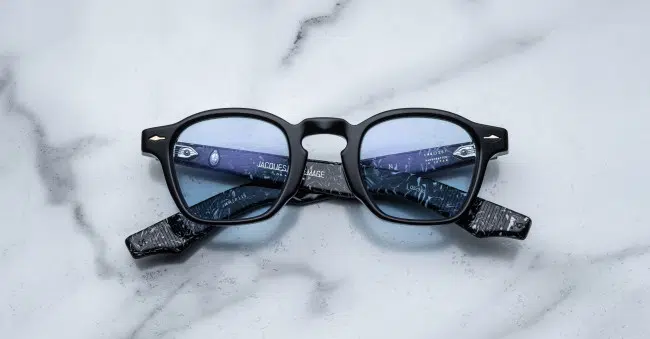 The Jacques Marie Mage Zephirin features black frames, blue-tinted lenses, and marbled black-patterned arms with text on the inside, shown displayed on a white marble surface.