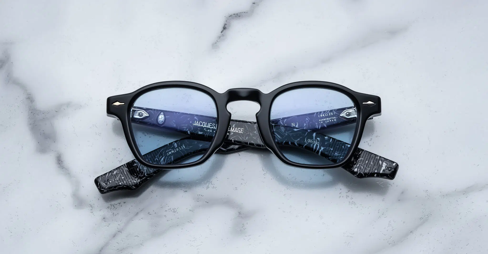 The Jacques Marie Mage Zephirin features black frames, blue-tinted lenses, and marbled black-patterned arms with text on the inside, shown displayed on a white marble surface.