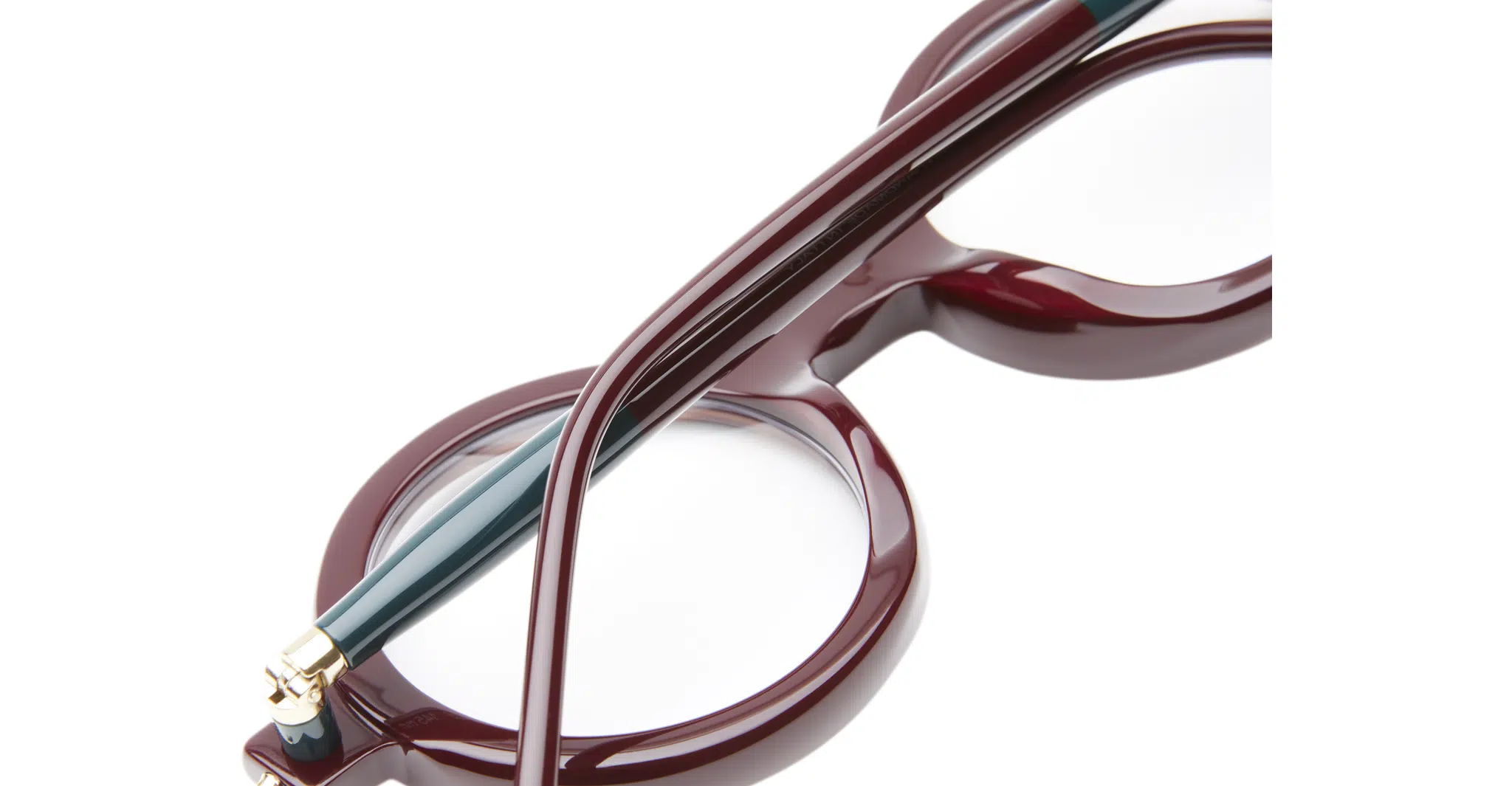 Kuboraum-P1-Bordeaux-KRO0P1DR000000OP-Detail2-Dan-Deutsch Close-up of Kuboraum P1 round eyeglasses with glossy burgundy frames and a green temple arm, set against a white background. Focus is on the frame's details and lenses.