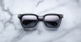 A pair of Jacques Marie Mage Borodino sunglasses with black square frames and dark lenses rests on a white marble surface.