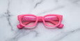 The Jacques Marie Mage Whiskeyclone features bright pink rectangular frames with slightly thick rims, displayed facing up on a white marble surface with subtle gray veins and inward-folded temples.