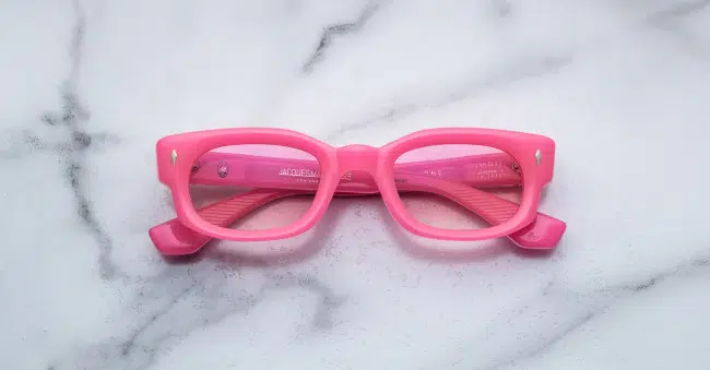 The Jacques Marie Mage Whiskeyclone features bright pink rectangular frames with slightly thick rims, displayed facing up on a white marble surface with subtle gray veins and inward-folded temples.