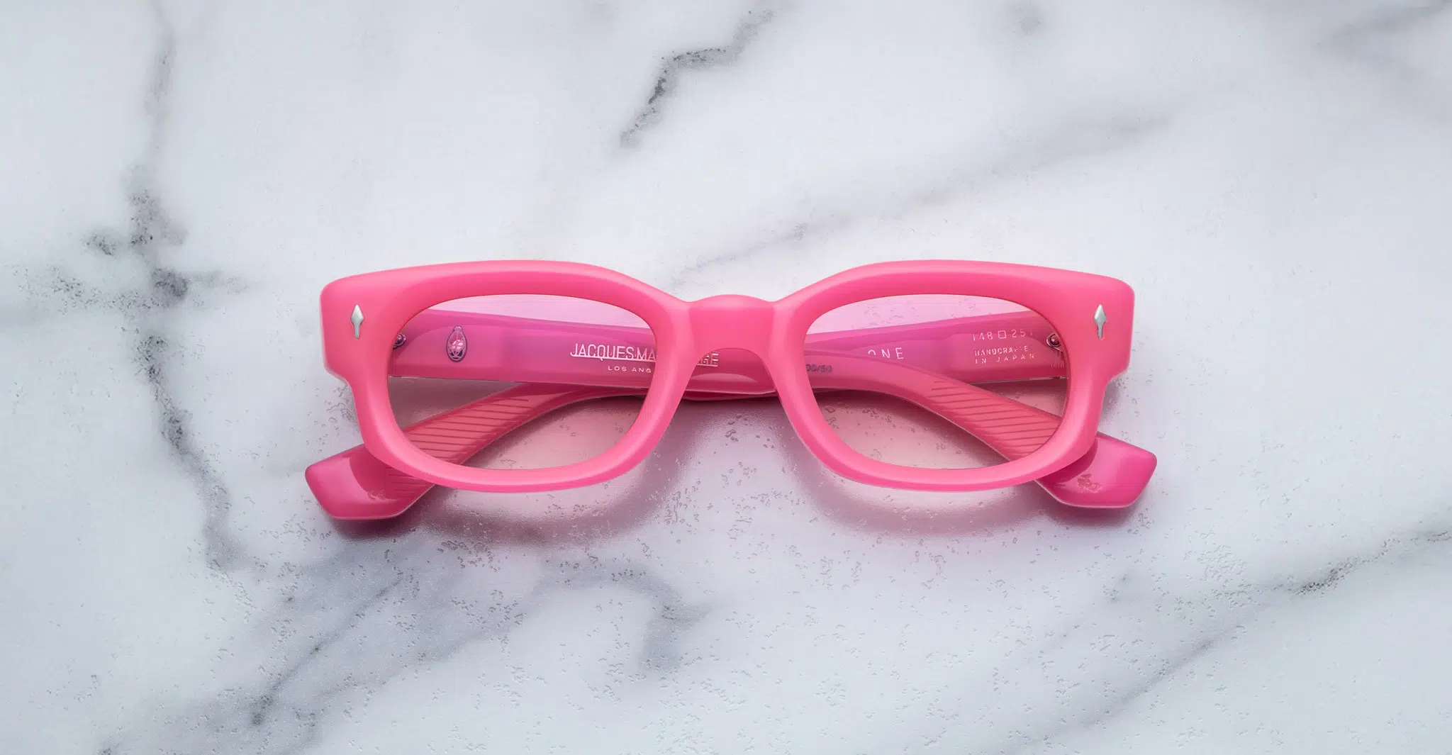 The Jacques Marie Mage Whiskeyclone features bright pink rectangular frames with slightly thick rims, displayed facing up on a white marble surface with subtle gray veins and inward-folded temples.