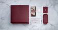 A large burgundy Jacques Marie Mage Windsor box is displayed on a marble surface with matching items: a folded cloth, card, small notebook, rectangular case, pen, and information card.