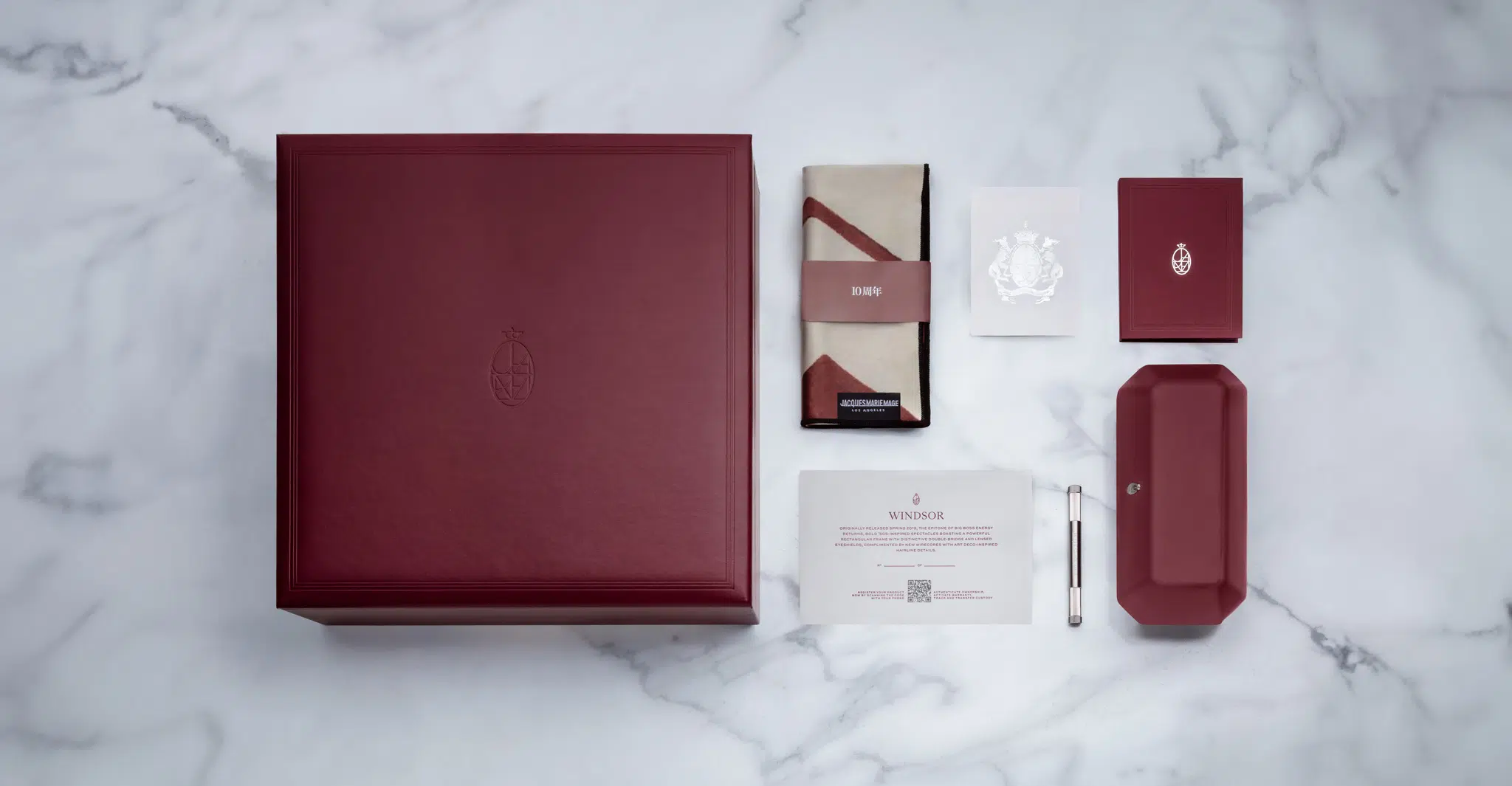 A large burgundy Jacques Marie Mage Windsor box is displayed on a marble surface with matching items: a folded cloth, card, small notebook, rectangular case, pen, and information card.