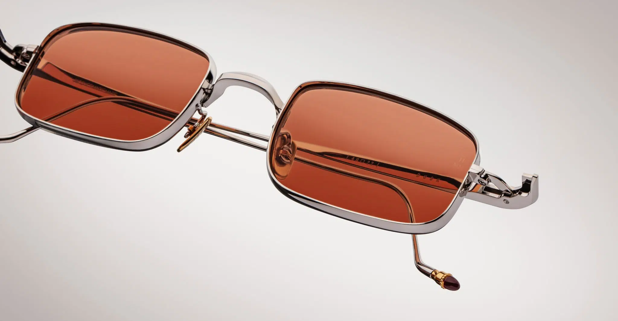 Jacques-Marie-Mage-Dusk-Silver-JMMSK-41-Detail2-Dan-Deutsch Close-up of Jacques Marie Mage Dusk sunglasses featuring rectangular metallic silver frames, orange-tinted lenses, and gold accents on the nose pads and temple tips, set against a light gradient background.