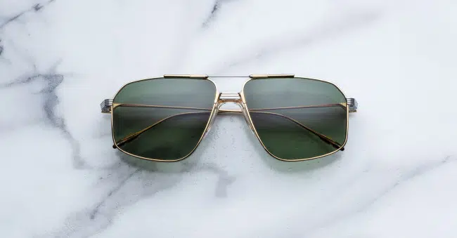 The Jacques Marie Mage Jagger sunglasses, featuring gold square frames and green lenses, rest on a white marble surface with gray veining.