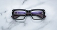 A pair of Jacques Marie Mage Walker black rectangular eyeglasses rests face up on a white marble surface, revealing purple inner arms with visible text and numbering through the lenses.