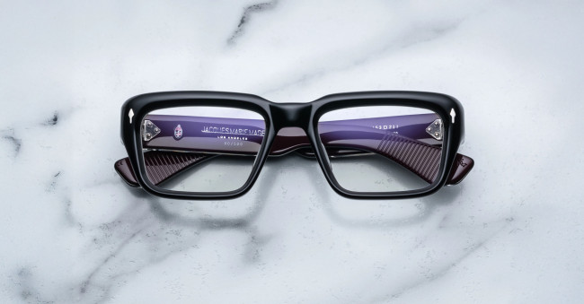 A pair of Jacques Marie Mage Walker black rectangular eyeglasses rests face up on a white marble surface, revealing purple inner arms with visible text and numbering through the lenses.