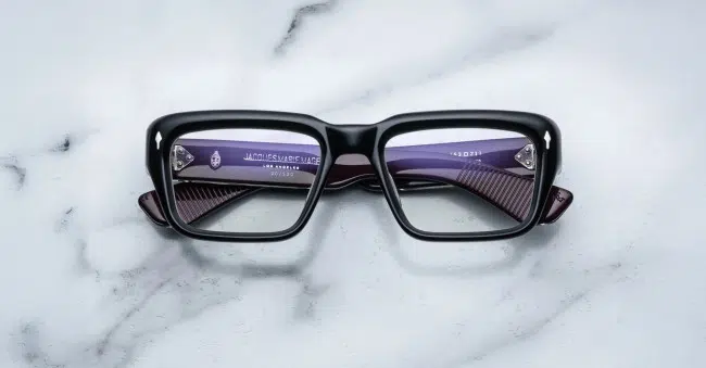 A pair of Jacques Marie Mage Walker black rectangular eyeglasses rests face up on a white marble surface, revealing purple inner arms with visible text and numbering through the lenses.
