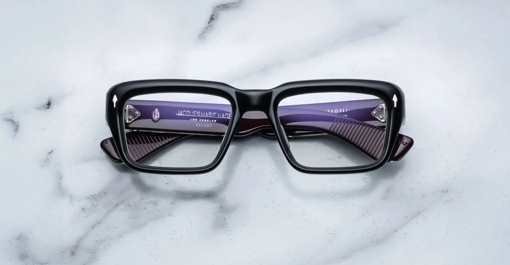 A pair of Jacques Marie Mage Walker black rectangular eyeglasses rests face up on a white marble surface, revealing purple inner arms with visible text and numbering through the lenses.