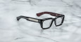 A pair of Jacques Marie Mage Walker rectangular black eyeglasses with thick frames and temples rests on a white marble surface. The inside of the right temple is burgundy and features white text.