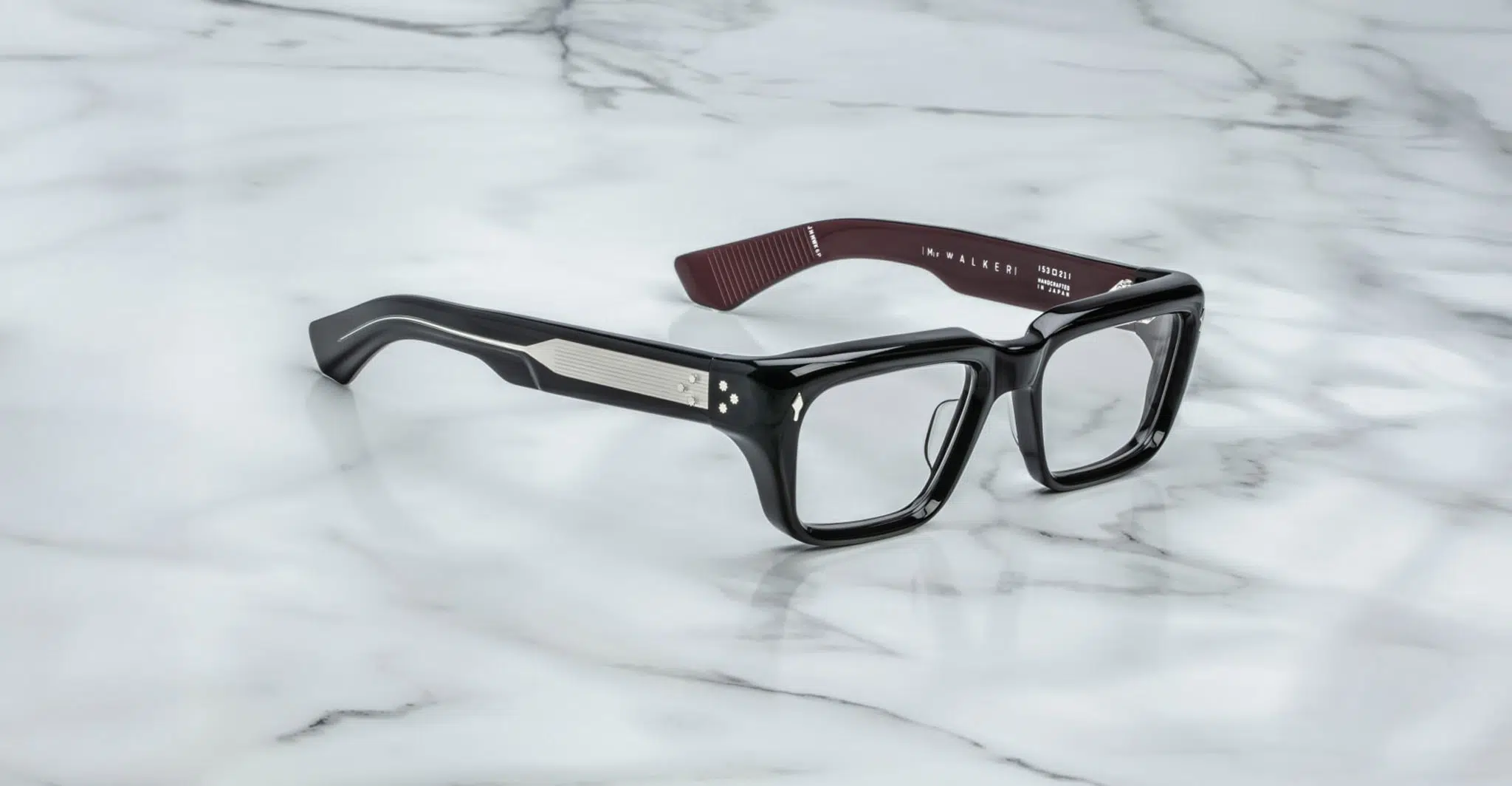 A pair of Jacques Marie Mage Walker rectangular black eyeglasses with thick frames and temples rests on a white marble surface. The inside of the right temple is burgundy and features white text.