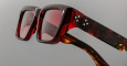 Close-up of Jacques Marie Mage Walker sunglasses with square, thick glossy red tortoiseshell frames, red-tinted lenses, and three small white star symbols on the arm by the hinge. The background is plain gray.