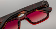 Close-up of Jacques Marie Mage Walker rectangular tortoiseshell sunglasses with red-tinted lenses, set against a neutral gray background. The photo highlights the distinctive frame details and bold lens color.