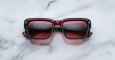 A pair of Jacques Marie Mage Walker rectangular, dark red sunglasses with tinted lenses rests on a white marble surface.