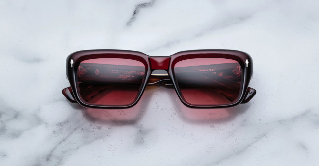 A pair of Jacques Marie Mage Walker rectangular, dark red sunglasses with tinted lenses rests on a white marble surface.