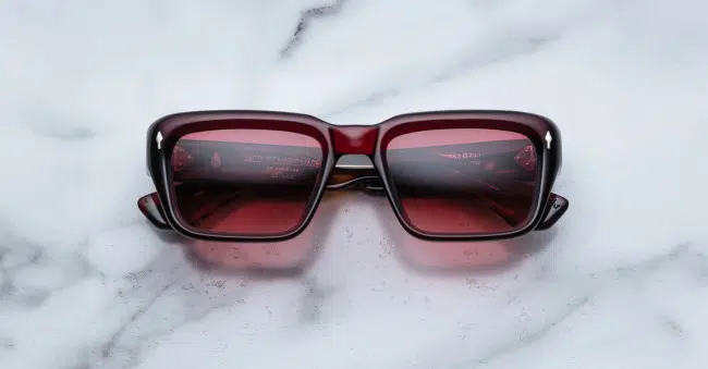 A pair of Jacques Marie Mage Walker rectangular, dark red sunglasses with tinted lenses rests on a white marble surface.