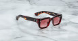 The Jacques Marie Mage Walker features square-shaped sunglasses with red tinted lenses and a thick tortoiseshell frame, displayed on a white marble surface.