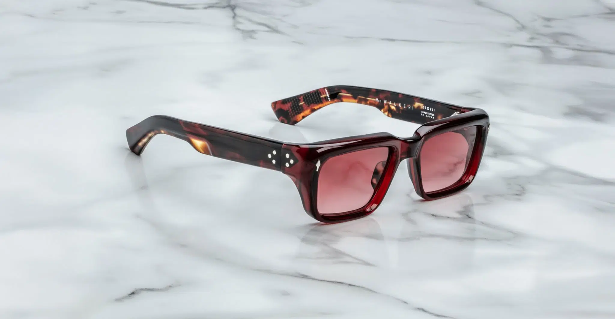 The Jacques Marie Mage Walker features square-shaped sunglasses with red tinted lenses and a thick tortoiseshell frame, displayed on a white marble surface.