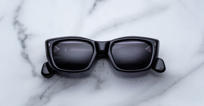A pair of Jacques Marie Mage Corniche black rectangular sunglasses with dark lenses rests on a white marble surface. The arms are partially open, revealing faint text on the inside of the lenses and frame.