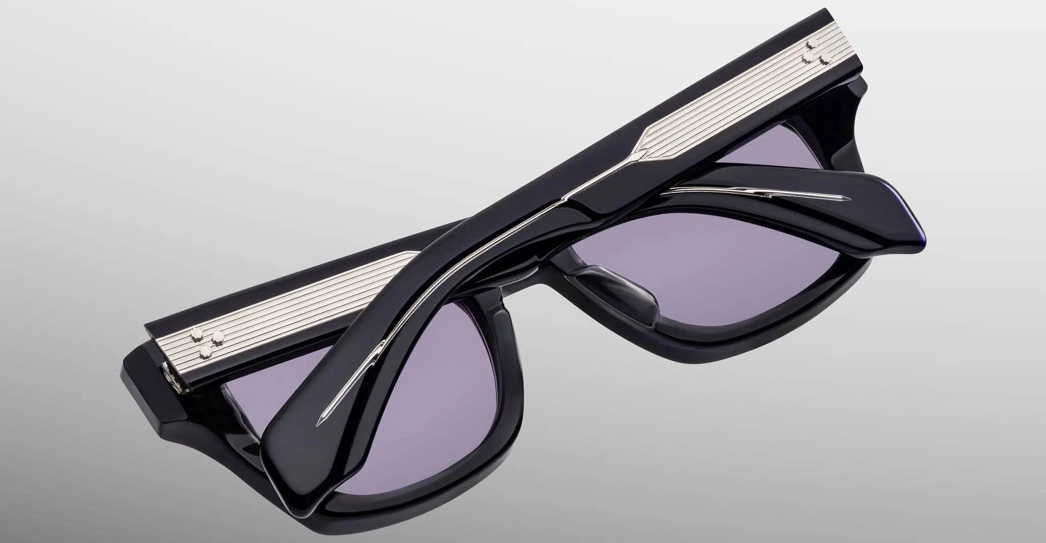 Jacques-Marie-Mage-Dealan-53-Hudson-JMMDEX-18U-Detail3-Dan-Deutsch The Jacques Marie Mage Dealan 53 is shown from above with temples crossed and folded, featuring black frames, silver accents, and purple-tinted lenses on a light gray background.
