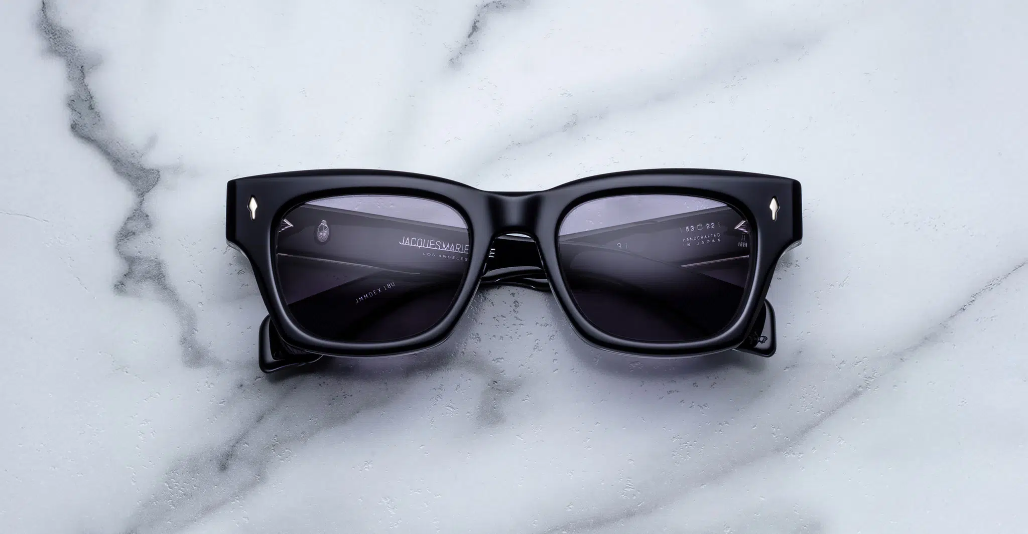 Jacques-Marie-Mage-Dealan-53-Hudson-JMMDEX-18U-Front-Dan-Deutsch The Jacques Marie Mage Dealan 53 sunglasses, featuring black rectangular thick frames, rest on a white marble surface with reflections visible on the dark lenses.